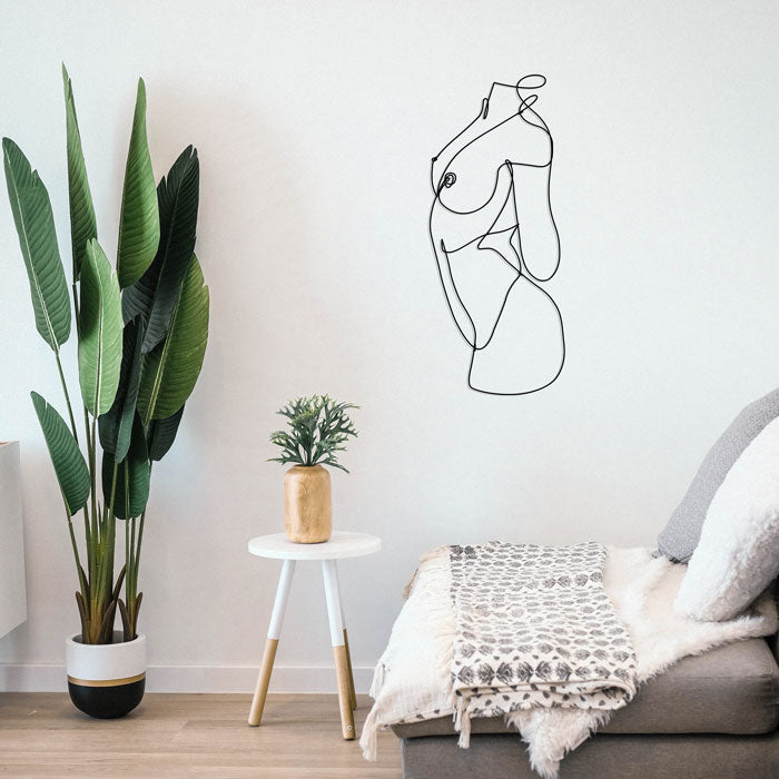 A modern living space features a large potted plant, a small round white table with a plant in a wooden pot, and a cushioned daybed with a patterned blanket. Above the bed, locally made hand-bent black steel wall art complements the minimalist line wall art of a female nude torso.