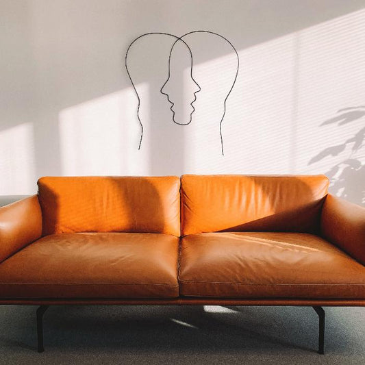 A minimalist living room showcases a modern brown leather couch, complemented by the abstract steel wall art from Living Things Creative hanging above it. This handcrafted piece overlapping profiles forming a central face, creating a visual illusion. Powder-coated in black for durability, with soft natural light casting dynamic shadows across the white wall.