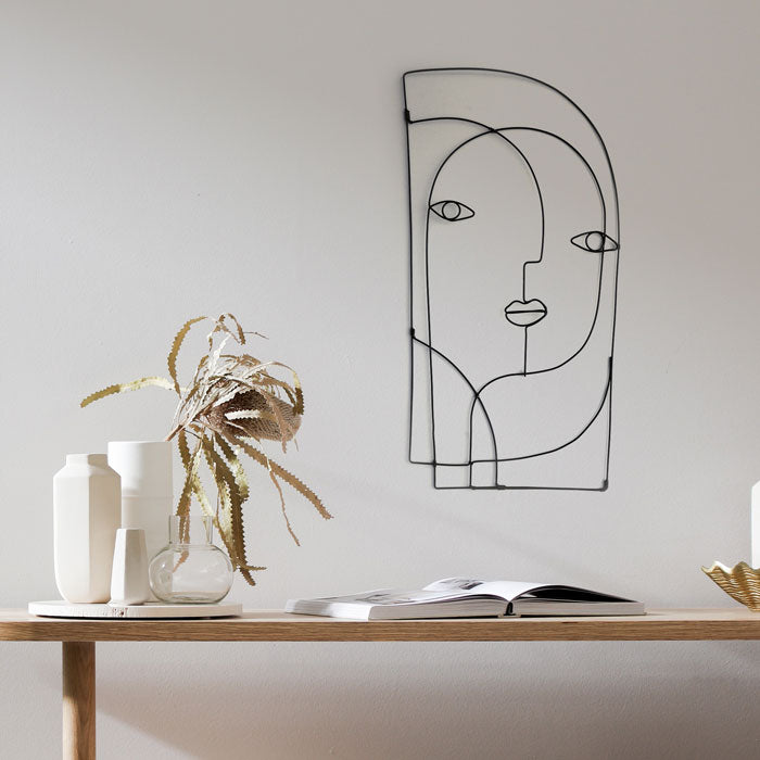 A minimalist room with a wooden table is adorned with white vases, a plant, and an open book. On the neutral-coloured wall hangs the "abstract steel wall art" by Living Things Creative in black, showcasing a depiction of an abstract face.