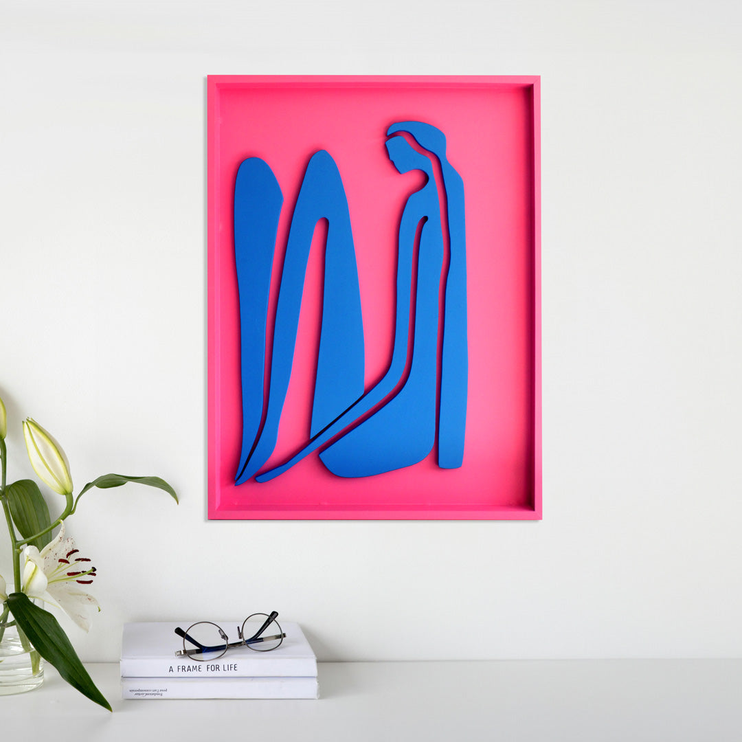 Handcrafted abstract wood wall art featuring blue organic shapes on a vibrant pink background, displayed above a minimalist table with books, eyeglasses, and a vase of white lilies