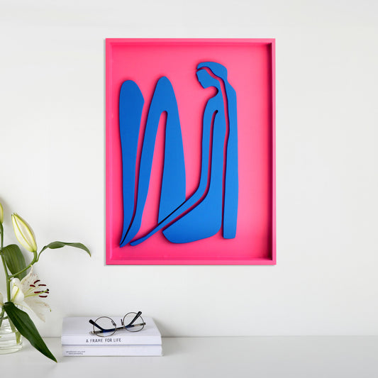 Handcrafted abstract wood wall art featuring blue organic shapes on a vibrant pink background, displayed above a minimalist table with books, eyeglasses, and a vase of white lilies