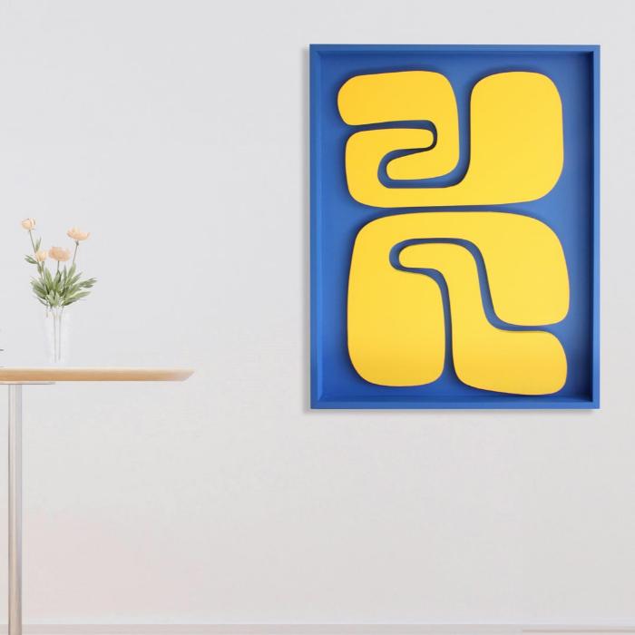 A minimalist interior includes a white armchair, a small table with two books and flowers, and the Origin wall art by living things creative. The piece features bright blue and yellow abstract forms, adding a textured design that lends depth to the space, resulting in an inviting yet serene atmosphere.