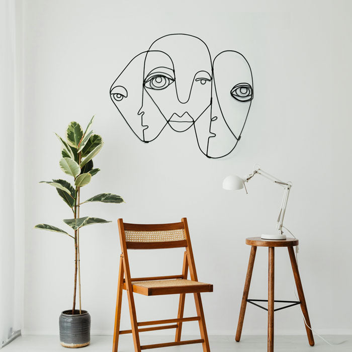 A minimalist room showcases a wooden chair and a tall potted plant next to a wooden stool adorned with a white desk lamp. A piece of black abstract steel wall art by Living Things Creative, depicting three faces, enhances the local wall art decor. Natural light pours in through a large window, accentuating every detail.