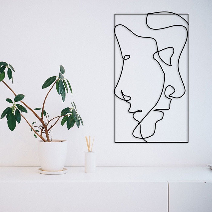 A minimalist room showcases a potted plant on a white table. The wall is adorned with a simple line art of a face, complemented by the abstract steel wall art from Living Things Creative in black. Additionally, the table features a small diffuser with reed sticks.